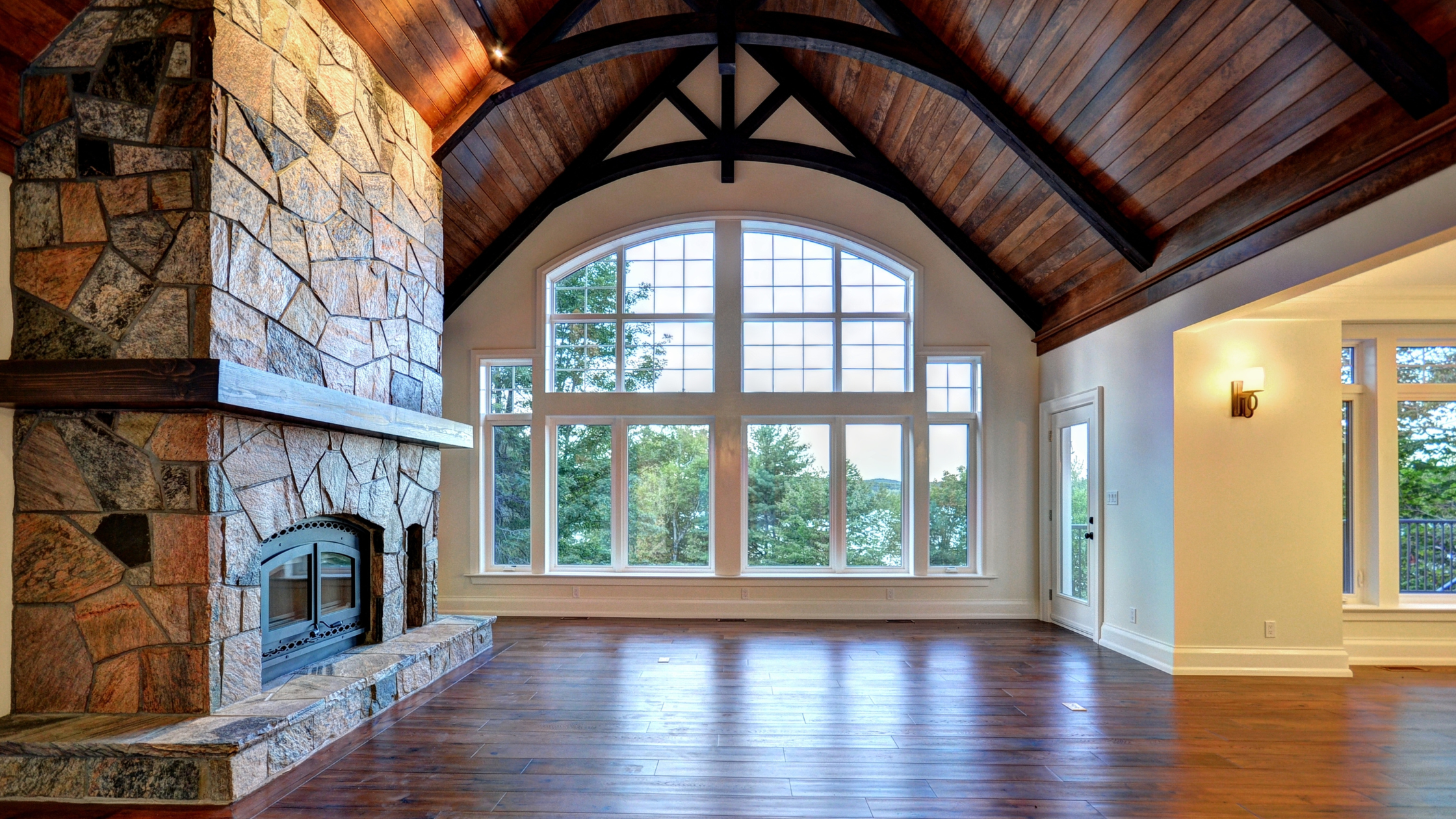 Luxury custom cottage interior in Muskoka featuring exposed timber beams, natural stone fireplace, and expansive arched windows overlooking lake views - showcasing premium finishes that impact the cost to build a custom cottage in Muskoka