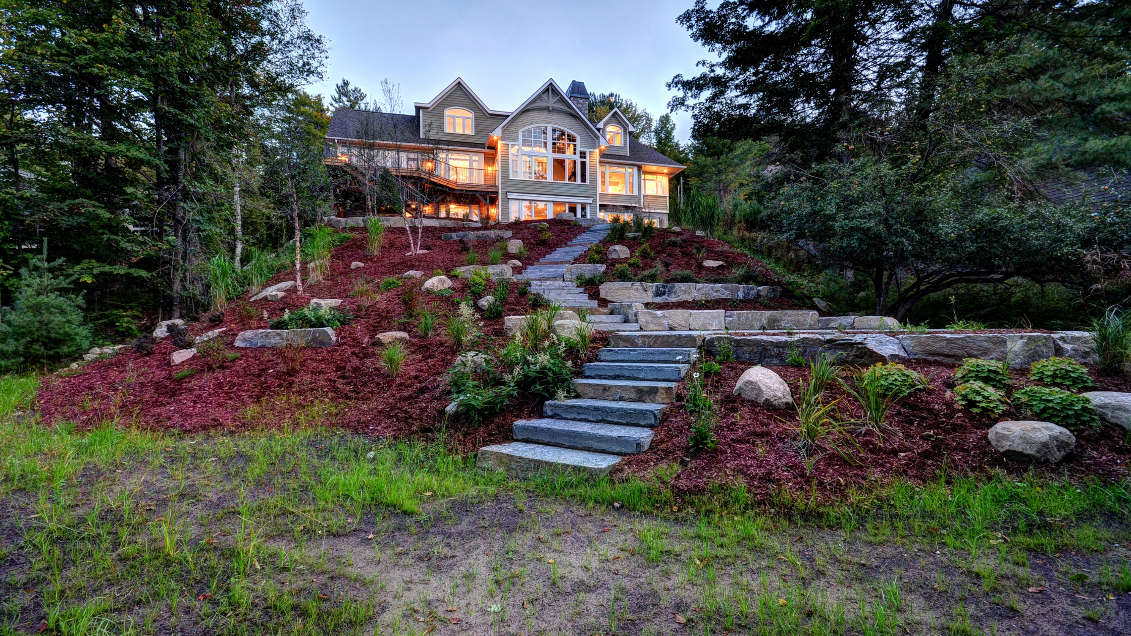 Custom cottage exterior in Muskoka built on rocky Canadian Shield terrain with extensive landscaping and stone retaining walls, demonstrating site preparation challenges that affect the cost to build a custom cottage in Muskoka