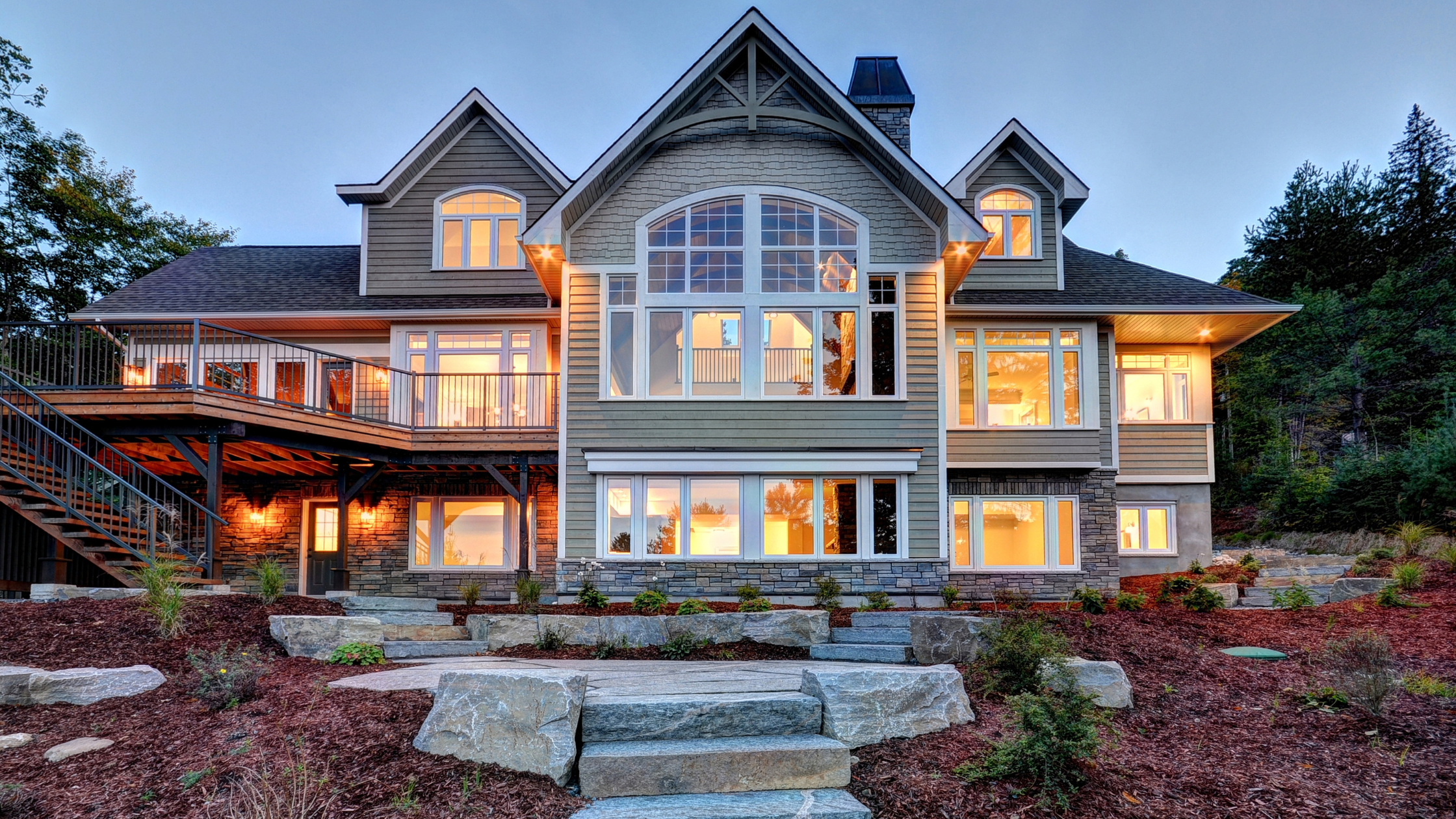 Luxury lakefront custom cottage in Muskoka featuring expansive windows, natural stone landscaping, and multi-level design with outdoor staircases, exemplifying high-end architectural elements that increase the cost to build a custom cottage in Muskoka